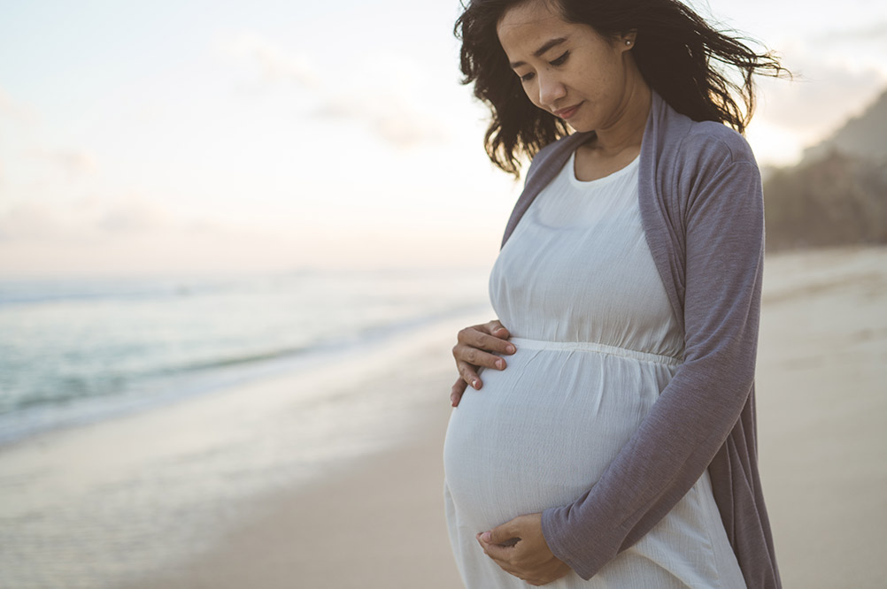 A pregnant woman holds her belly while standing on the shoreline