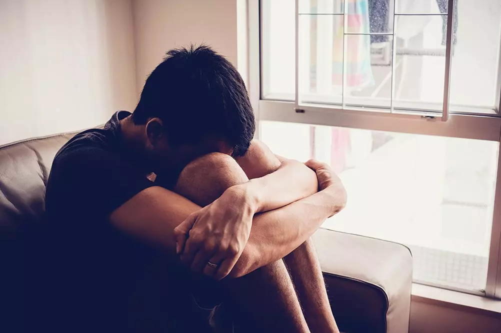 A young male experiencing PTSD or post-traumatic stress disorder hugs his knees on a couch