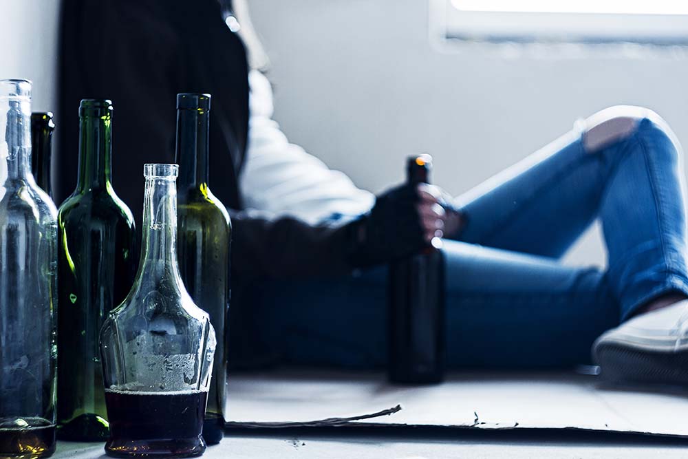 A man struggling with alcohol addiction sits on the ground next to liquor and wine bottles