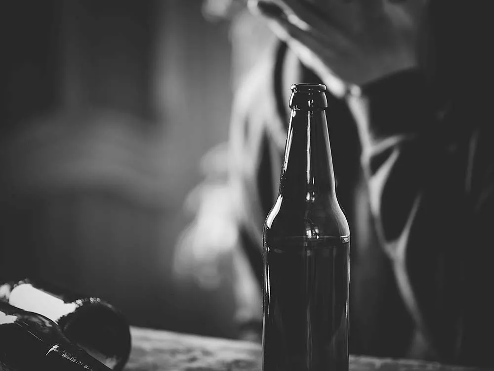 Black and white image with a beer bottle in the foreground and someone struggling with alcohol addiction in the background