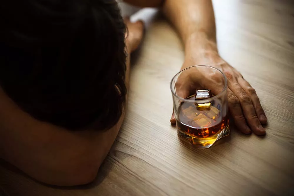 A man with his head odwn on a table holding a glass of alcohol