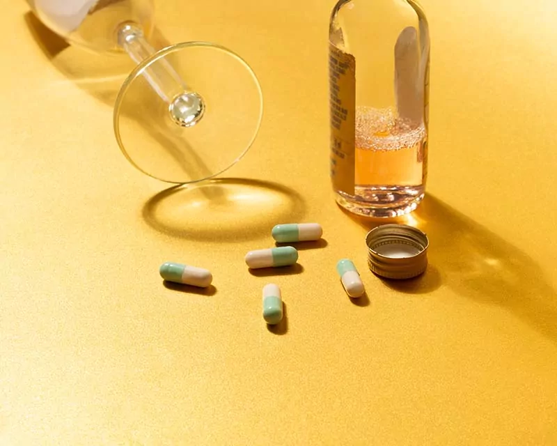 Lexapro pills next to a small bottle of alcohol and an overturned glass on a yellow background