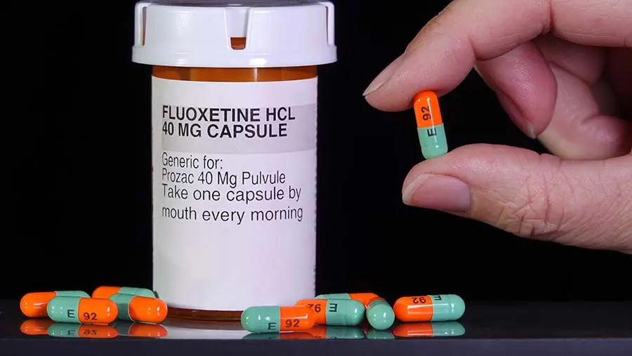 A hand holds a Prozac or Fluoxetine pill next to a pill bottle and pills scattered on a table