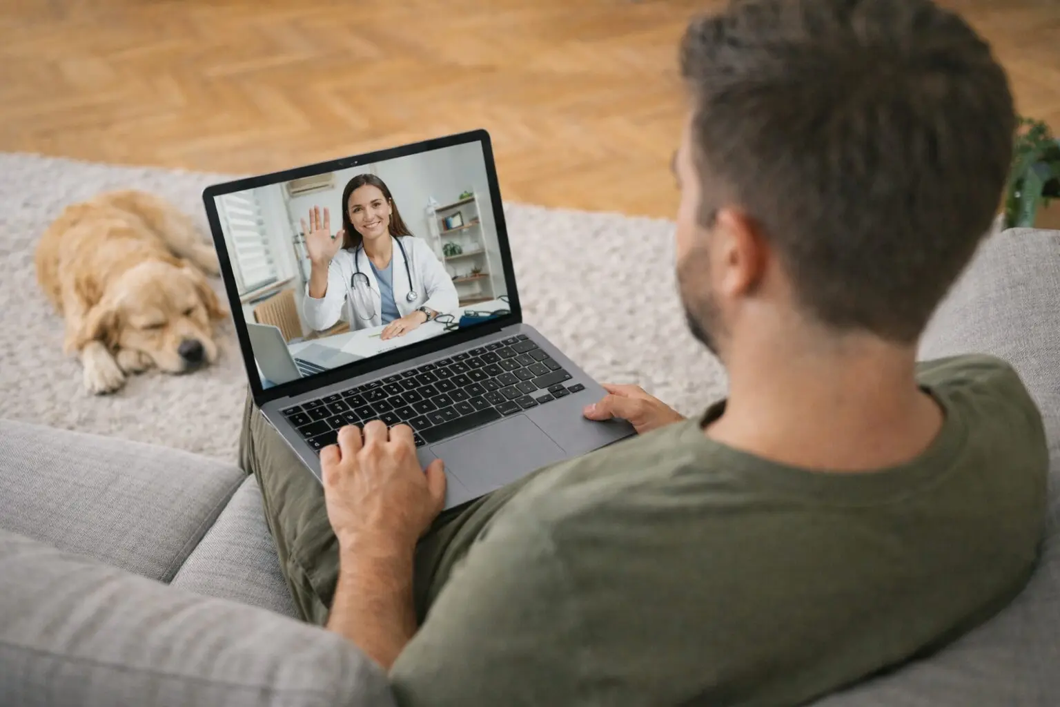 A man is sitting on a cozy couch using his laptop computer to talking with a provider during a telehealth appointment.