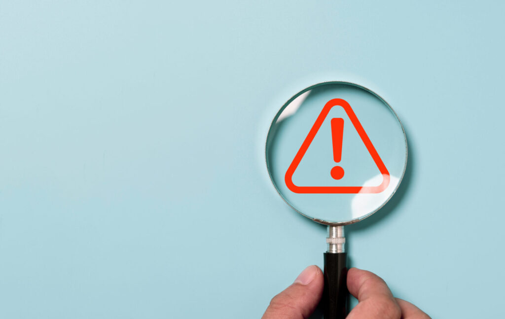 Hand holding magnifier glass with red triangle caution warning sign for focus notification error and maintenance concept - Eleanor Health Hand holding a magnifying glass showing a red warning triangle with an exclamation mark.